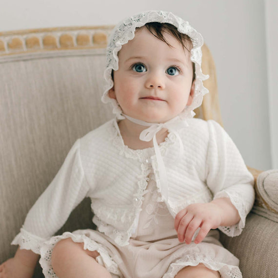 A baby with big blue eyes sits on a beige chair, wearing the Charlotte Quilted Cotton Sweater over the Charlotte Romper and Lace Bonnet. The sweater is adorned with rose and vine lace trim. The baby looks slightly upward, with one hand resting on the armrest.