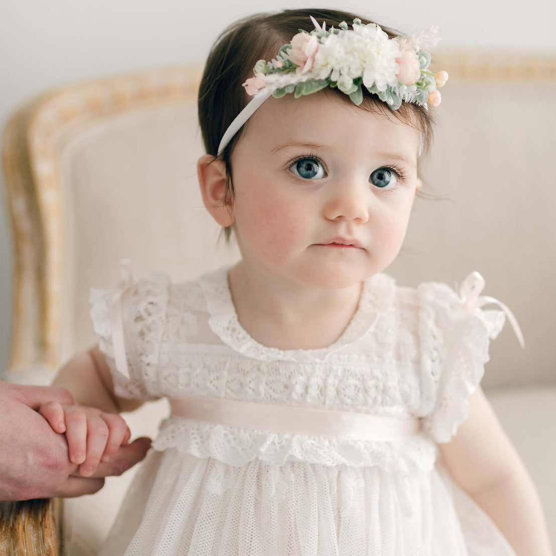 Baby girl sitting on a linen chair, wearing the Charlotte Flower Headband with soft pink and white flowers. Her large blue eyes gaze ahead as she holds onto someone's hand.