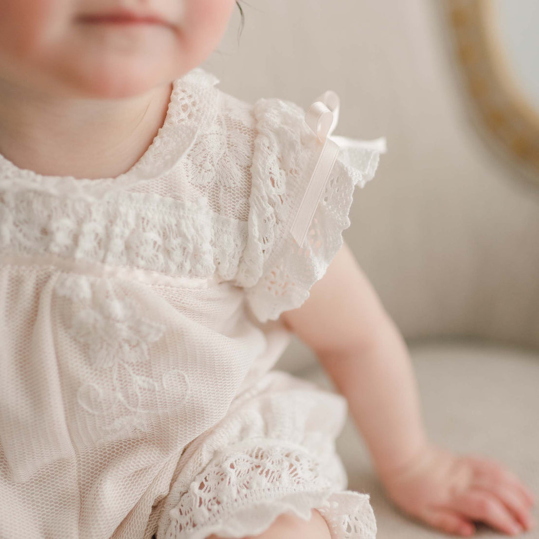 Close-up of a baby girl wearing the Charlotte Romper with a ribbon on the shoulder. The photo focuses on the shoulder and short sleeve of the romper. The baby is sitting on a light-colored chair.