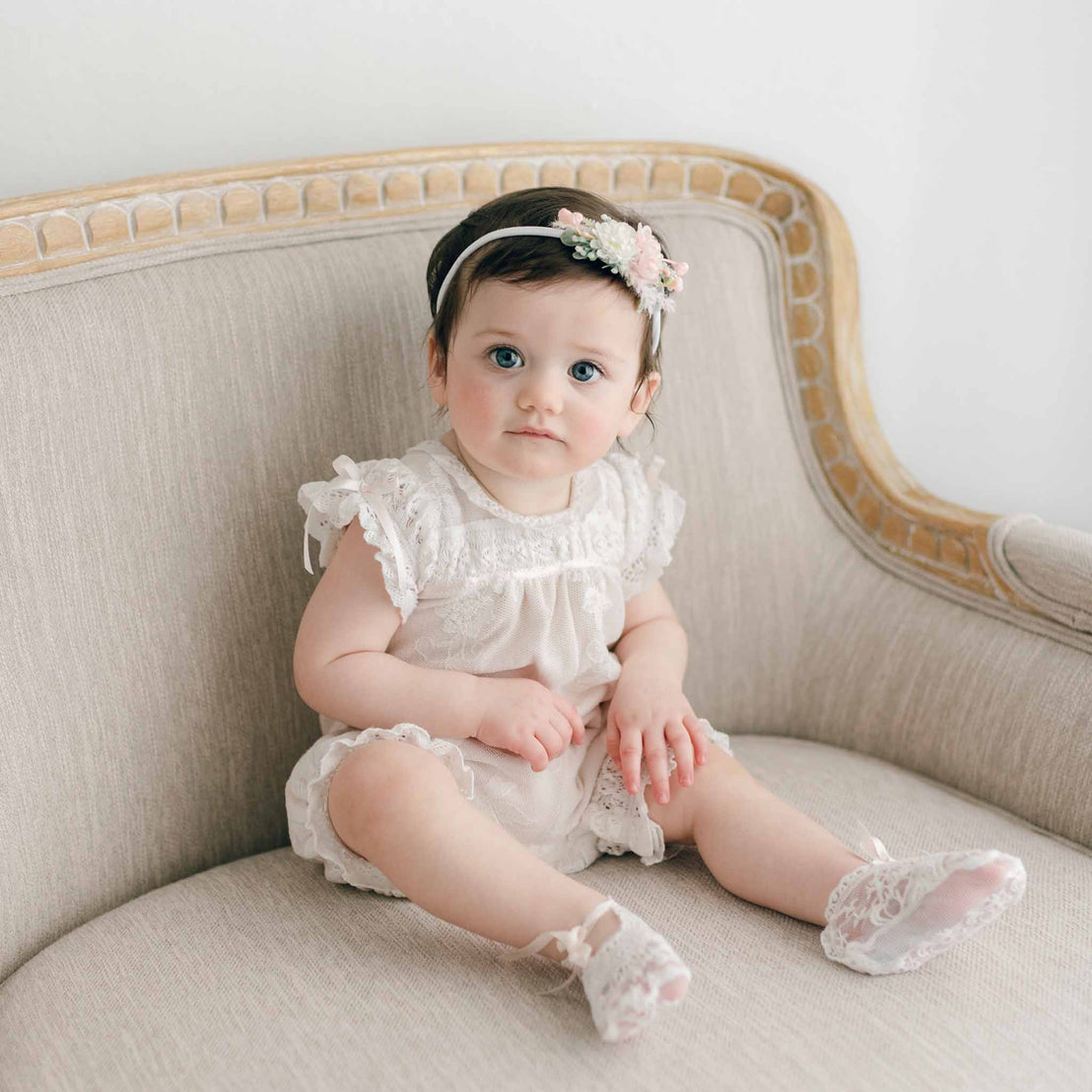 A baby girl with dark hair, sitting on a beige cushioned sofa. She is wearing a Charlotte Romper and matching lace booties, with a floral headband adorning her head. The background is plain white, highlighting the baby and the sofa.