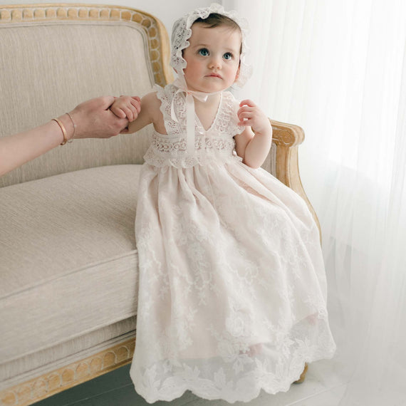 A baby girl in a Charlotte Christening Gown & Bonnet sits on an elegant chair, looking curiously towards the camera. Her head is adorned with a matching lace bonnet.