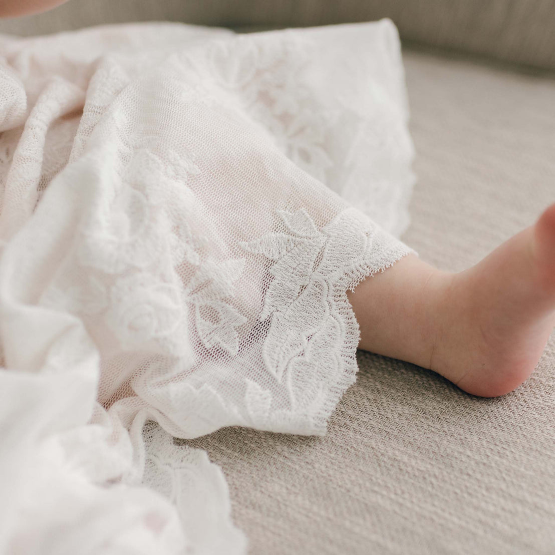 The skirt of the Charlotte Christening Gown with cotton lace scallop hem featured on a baby girl sitting on a beige chair.