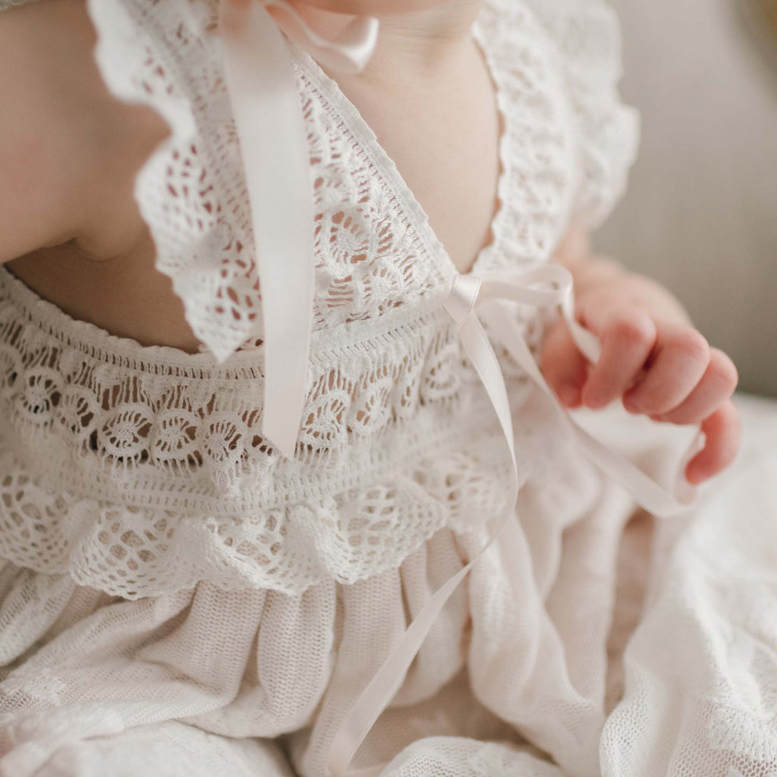 Close-up of a delicate Charlotte Christening Gown with intricate cotton lace details and a soft pink ribbon, displayed on a hanger. The texture of the fabric showcases a fine, detailed pattern.