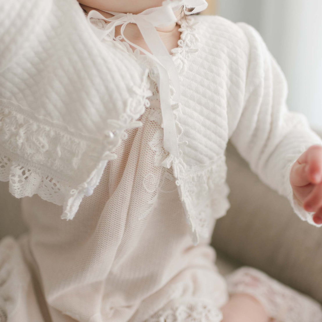 A close up of the Charlotte Quilted Cotton Sweater. A baby girl is wearing the sweater over the Charlotte Romper. The Charlotte Quilted Cotton Sweater features intricate rose and vine lace details.