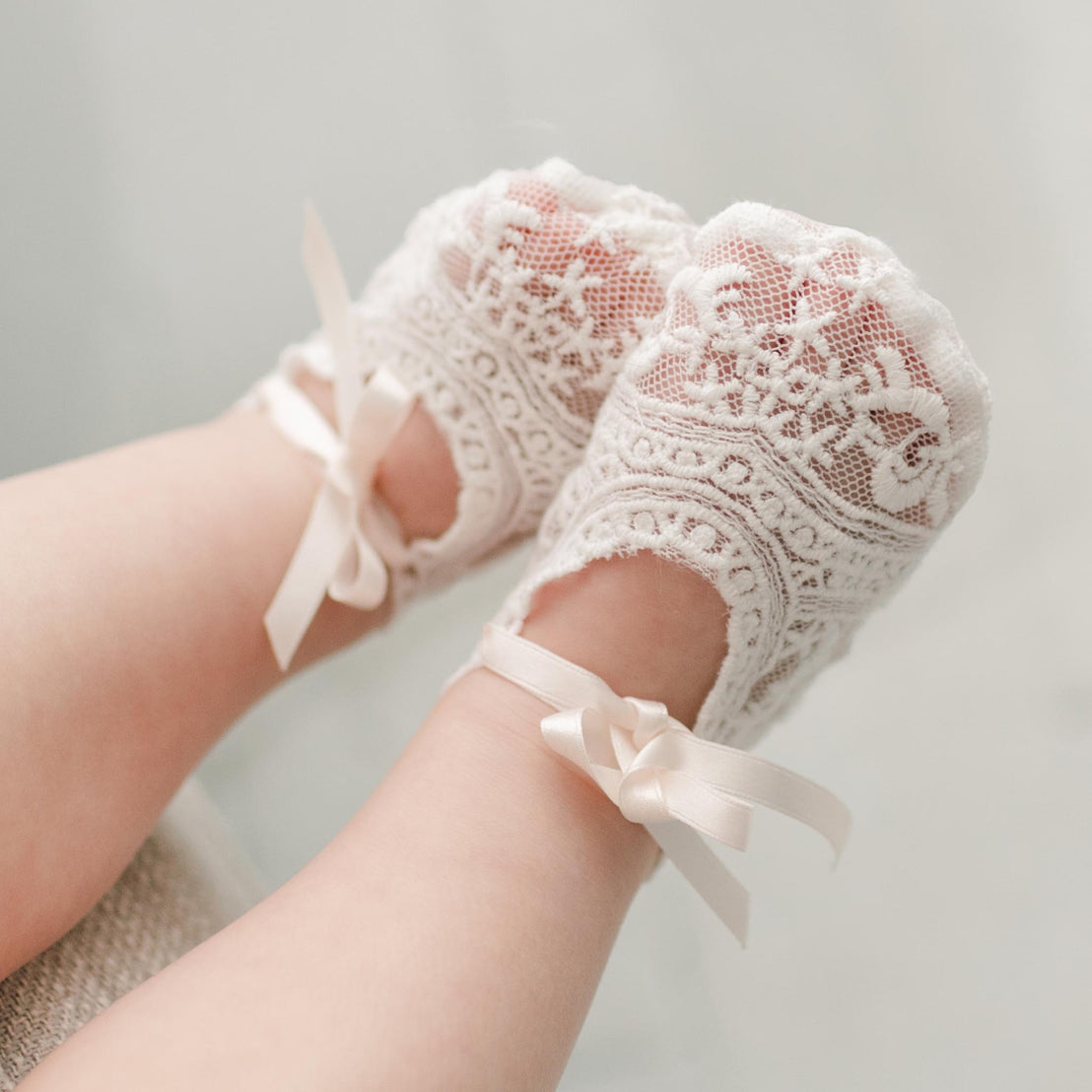 Close-up of a baby's feet wearing the Charlotte Booties made of light ivory lace, adorned with intricate patterns and tied with pink silk ribbon ties. The background is softly blurred, emphasizing the intricate details of the booties.