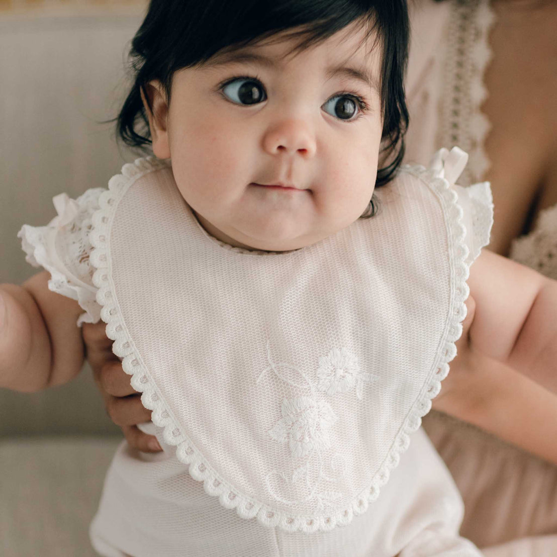 A baby with dark hair and large eyes wears the Charlotte Bib with lace trim and floral embroidery. The baby is being held by an adult, only partially visible in the background. The baby’s expression is curious and slightly amused.