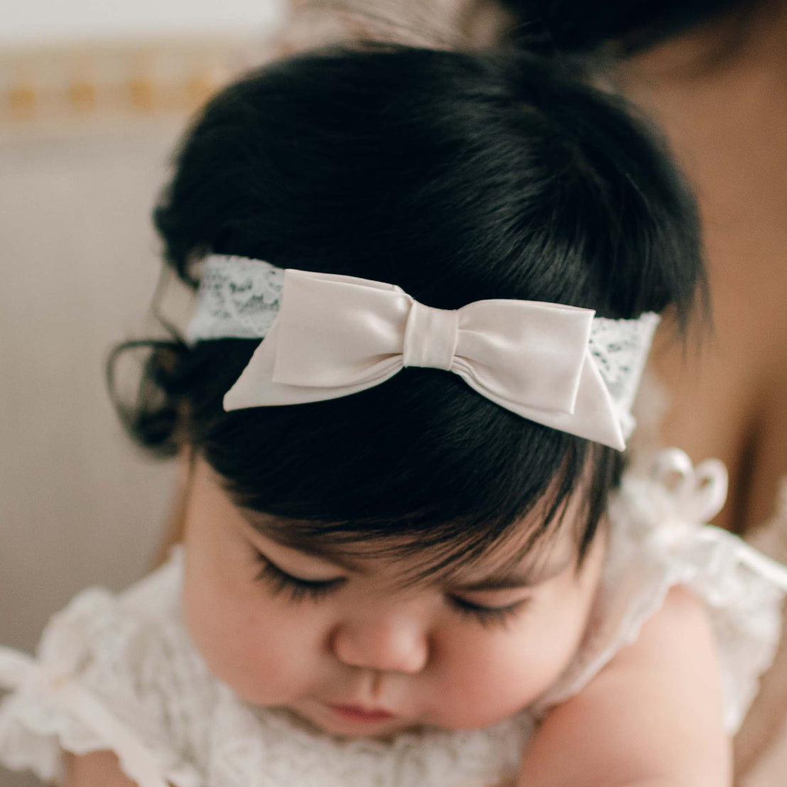 A baby girl with dark hair wearing the Charlotte Lace Headband. The headband features a pink champagne silk bow and stretch floral lace. The baby is looking down area from the camera.