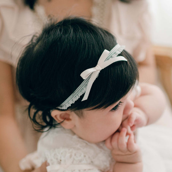 Close-up of a baby girl wearing the Charlotte Pink Silk Ribbon Headband, featuring delicate lace and a soft pink silk bow, while seated on her mother's lap.