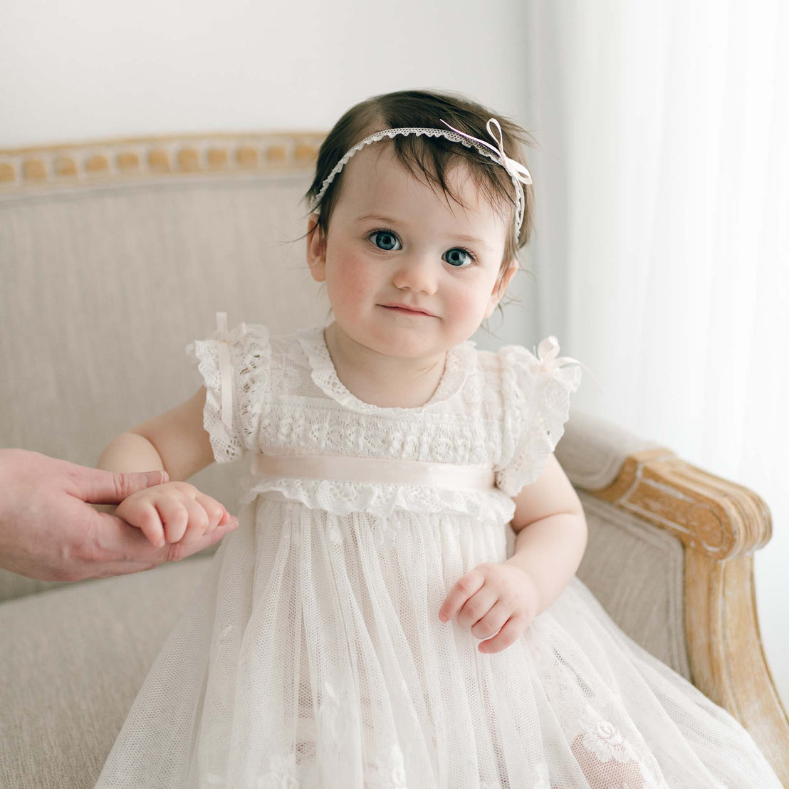 A baby wearing the Charlotte Convertible Skirt & Romper Set, complete with embroidered cotton lace and a delicate headband, is sitting on an upholstered chair. The baby holds the hand of an adult who is off to the side. The setting is softly lit and exudes a serene, airy feel.