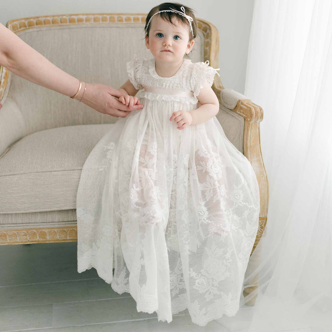 A baby girl in the Charlotte Convertible Skirt & Romper Set sits on a cream-colored armchair, adorned with a matching headband and gazing slightly to the side while holding an adult's hand. The background features sheer white curtains, enhancing the soft ambiance of the scene.