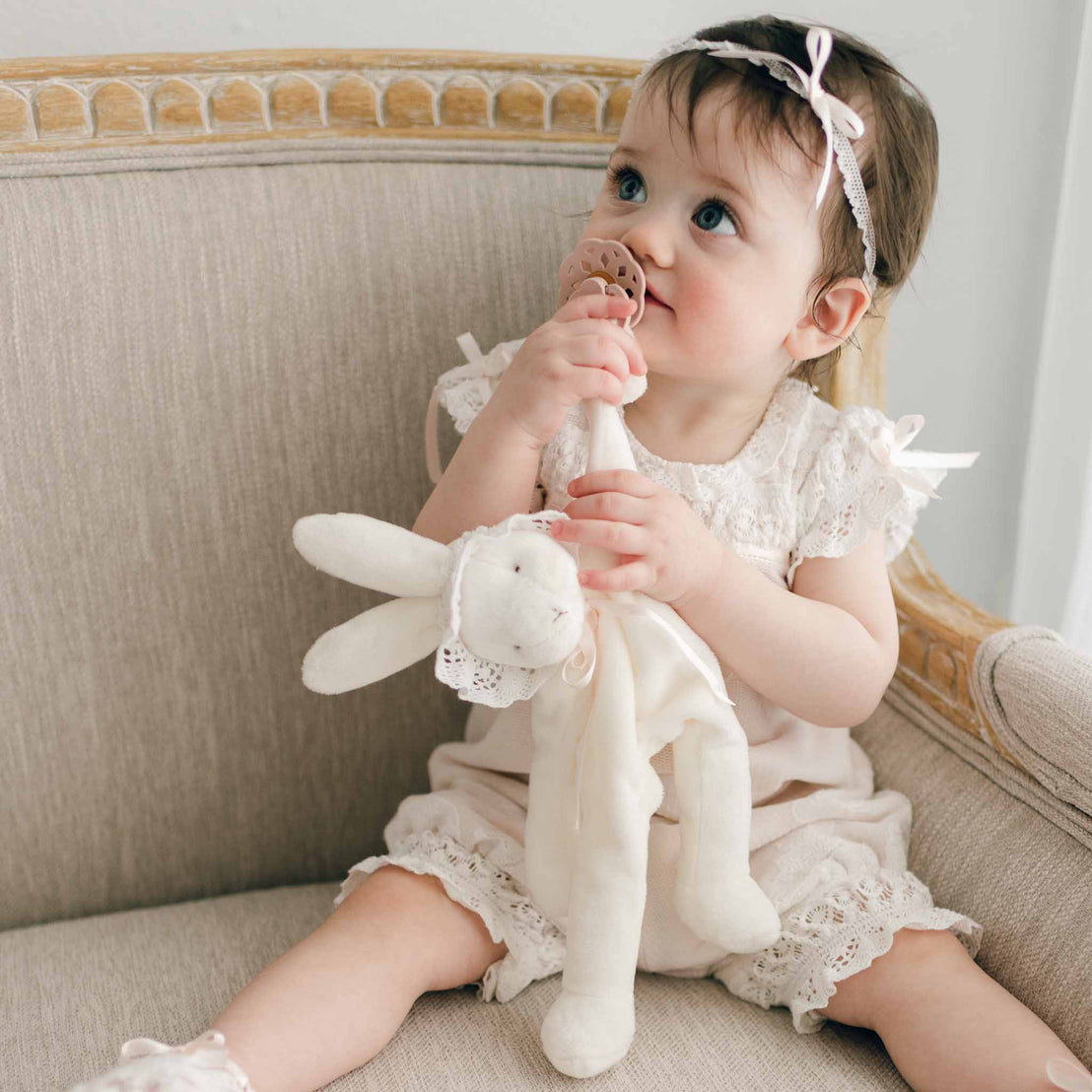 A young girl wearing the Charlotte Romper sits on a cushioned chair, holding a Charlotte Silly Bunny Buddy and Pacifier Holder to her mouth.