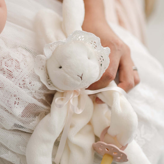 A close-up of a soft, white Charlotte Silly Bunny Buddy and Pacifier Holder dressed in a lace bonnet and ribbon. The bunny is being gently held by one hand. The edge of lace fabric and a soft pink pacifier clip are visible in the bunny's hand..