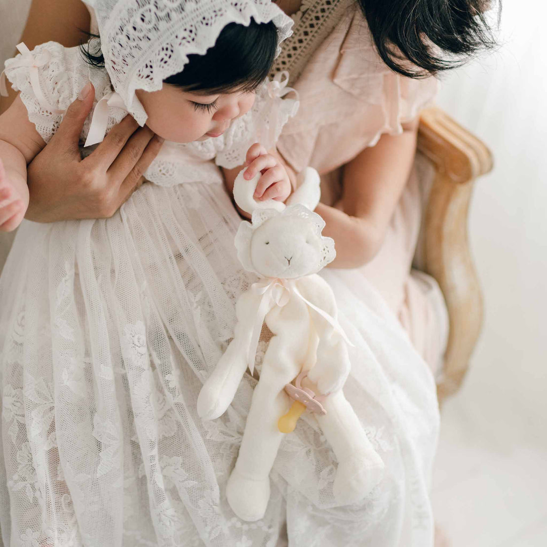 A baby in the Charlotte Charlotte Convertible Skirt & Romper Set and Bonnet is being held by her mother. The baby girl clutches a Charlotte Silly Bunny Buddy and Pacifier Holder.