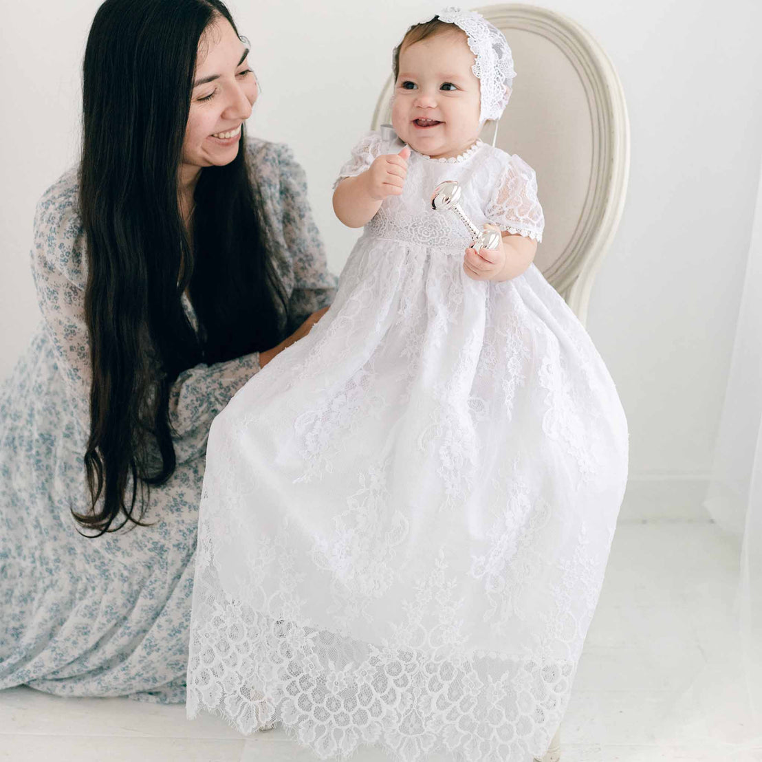 A joyful mother sits next to her baby dressed in a Olivia Christening Gown & Bonnet for a christening. The baby, holding a small rattle and smiling broadly.