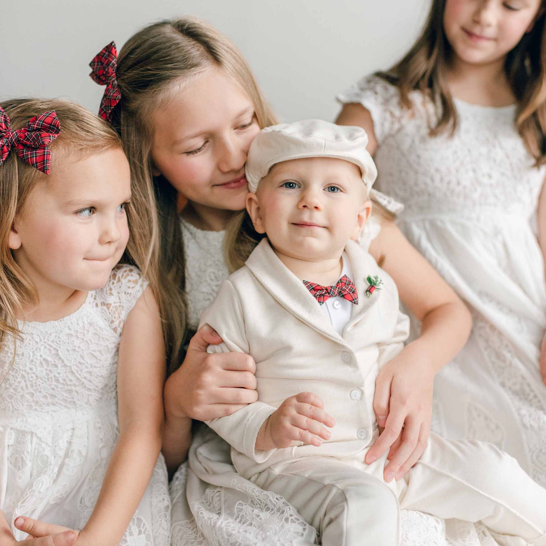 Three young girls in Grace Girls Lace Holiday Dresses, made from a luxurious silk and cotton blend, are gathered together with a baby boy wearing a cream outfit and cap. The girls have red bows in their hair and gently smile at the baby, creating a warm and tender atmosphere.