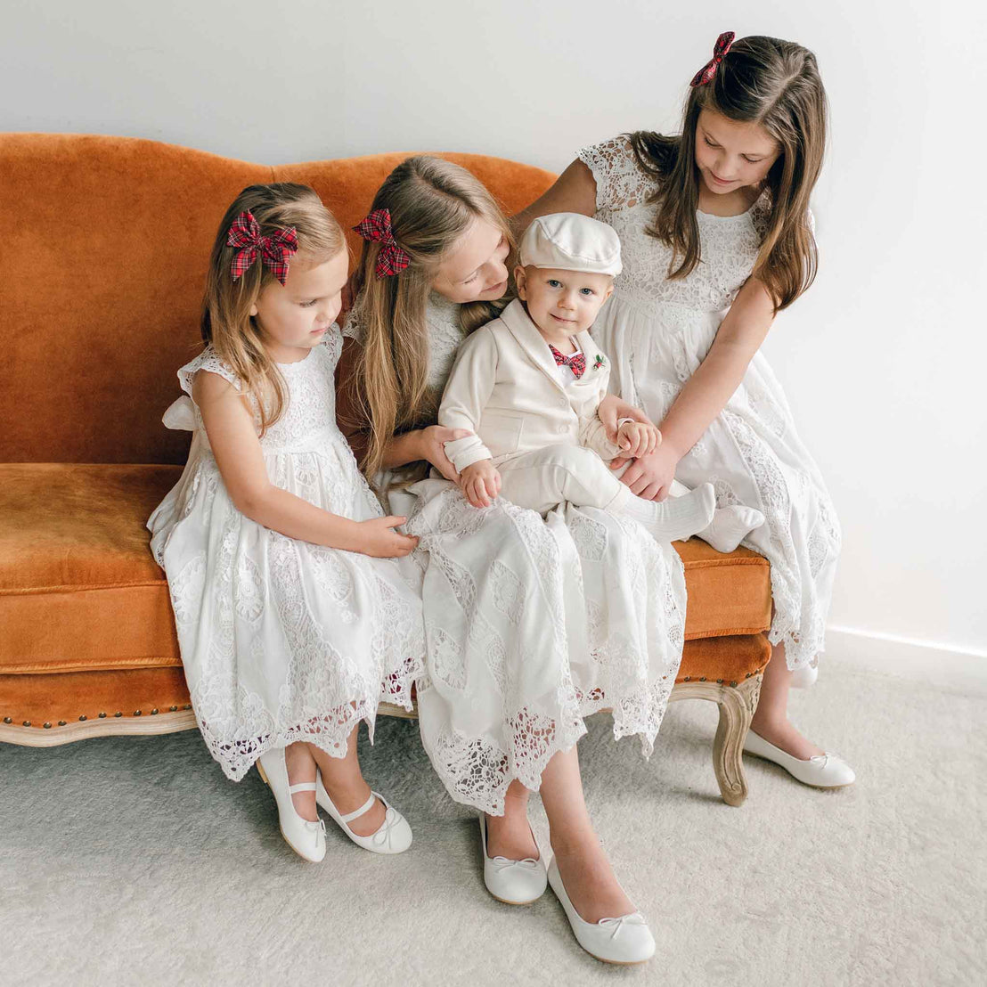 A group of four children in white dress clothes sits on an orange sofa. The girls, wearing Grace Girls Lace Holiday Dresses with tea-length style and red hair bows, are gathered around a boy dressed in a white suit and cap. They seem engaged and content together.