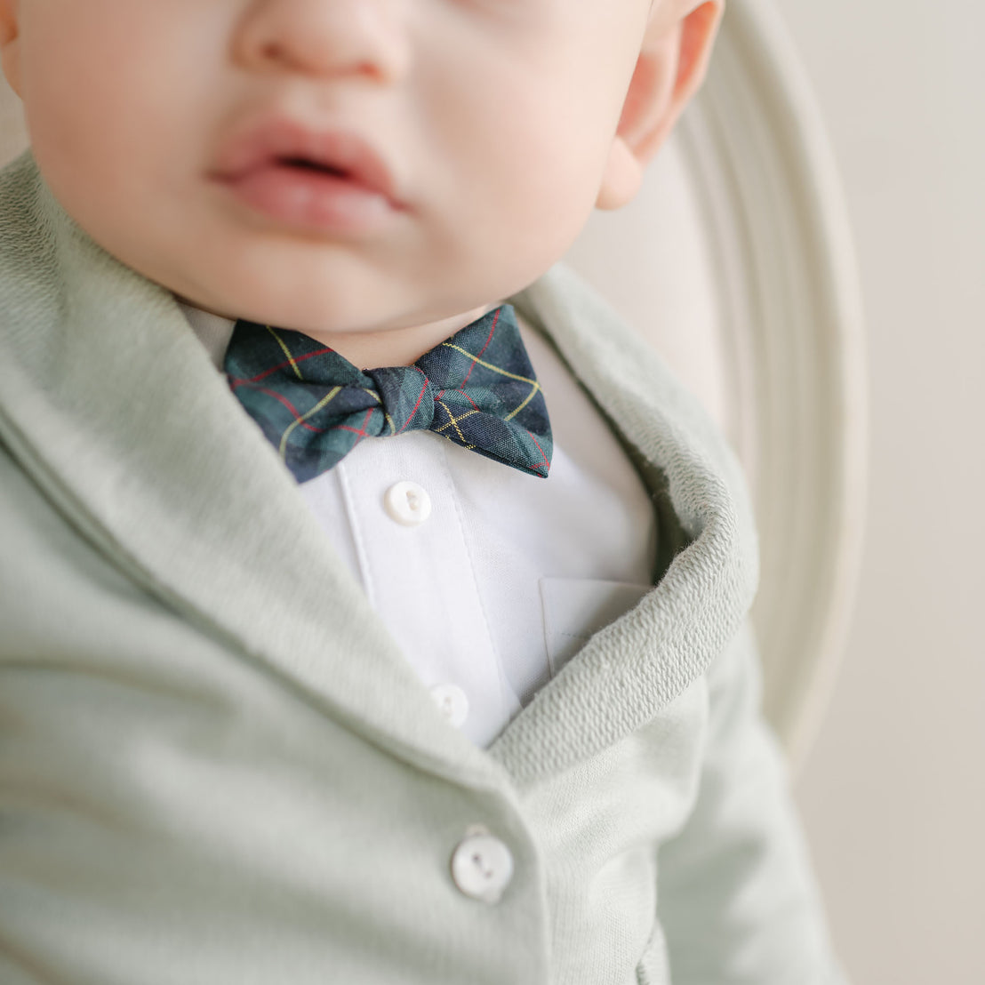 A close-up of a baby wearing the Cole 3-Piece Suit, featuring a green sweater crafted from soft French Terry cotton layered over a white shirt, complemented by a dark striped bow tie. This delightful ensemble is perfect for any holiday party. The focus is on the outfit and mouth against a light, neutral background.