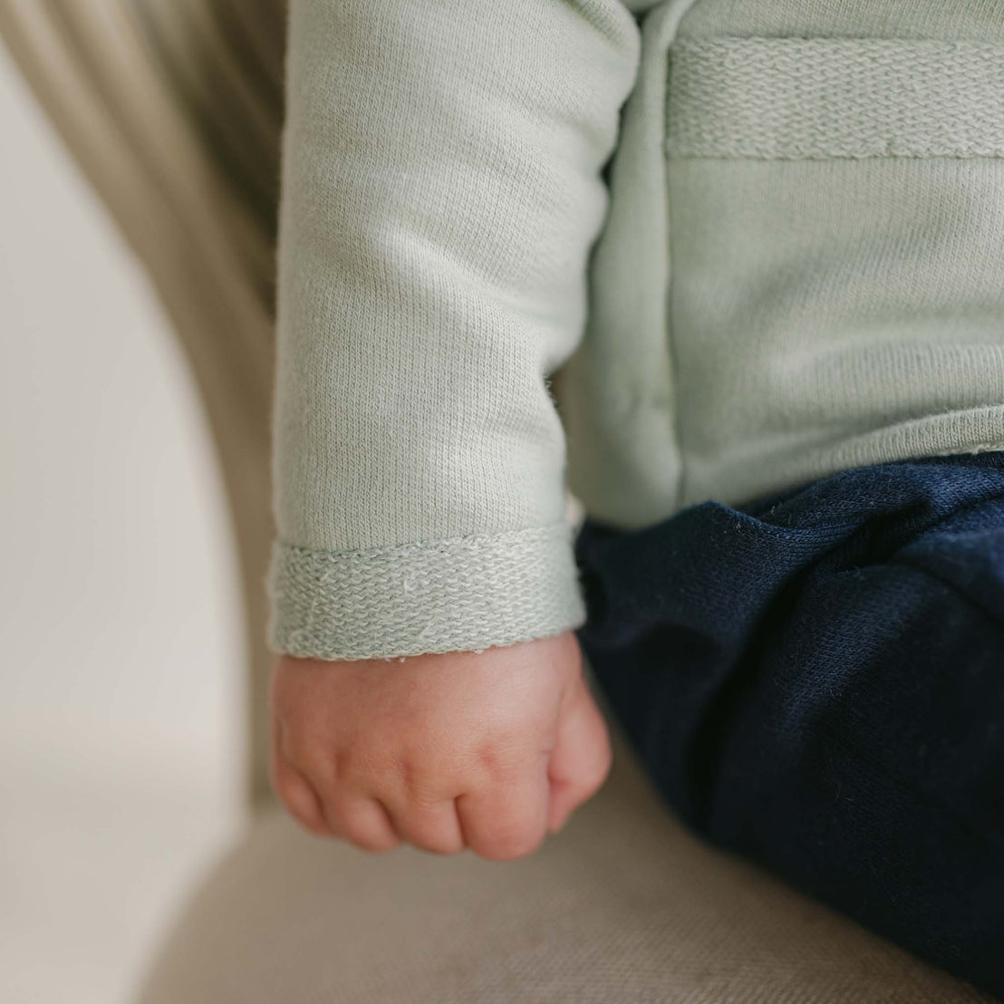 Close-up of a baby's hand resting on their lap, wrapped in the cozy embrace of the Cole 3-Piece Suit's light green French Terry cotton sweater and dark pants. Sitting on a beige chair, the focus remains on their delicate hand and the exquisite texture of this handmade clothing.