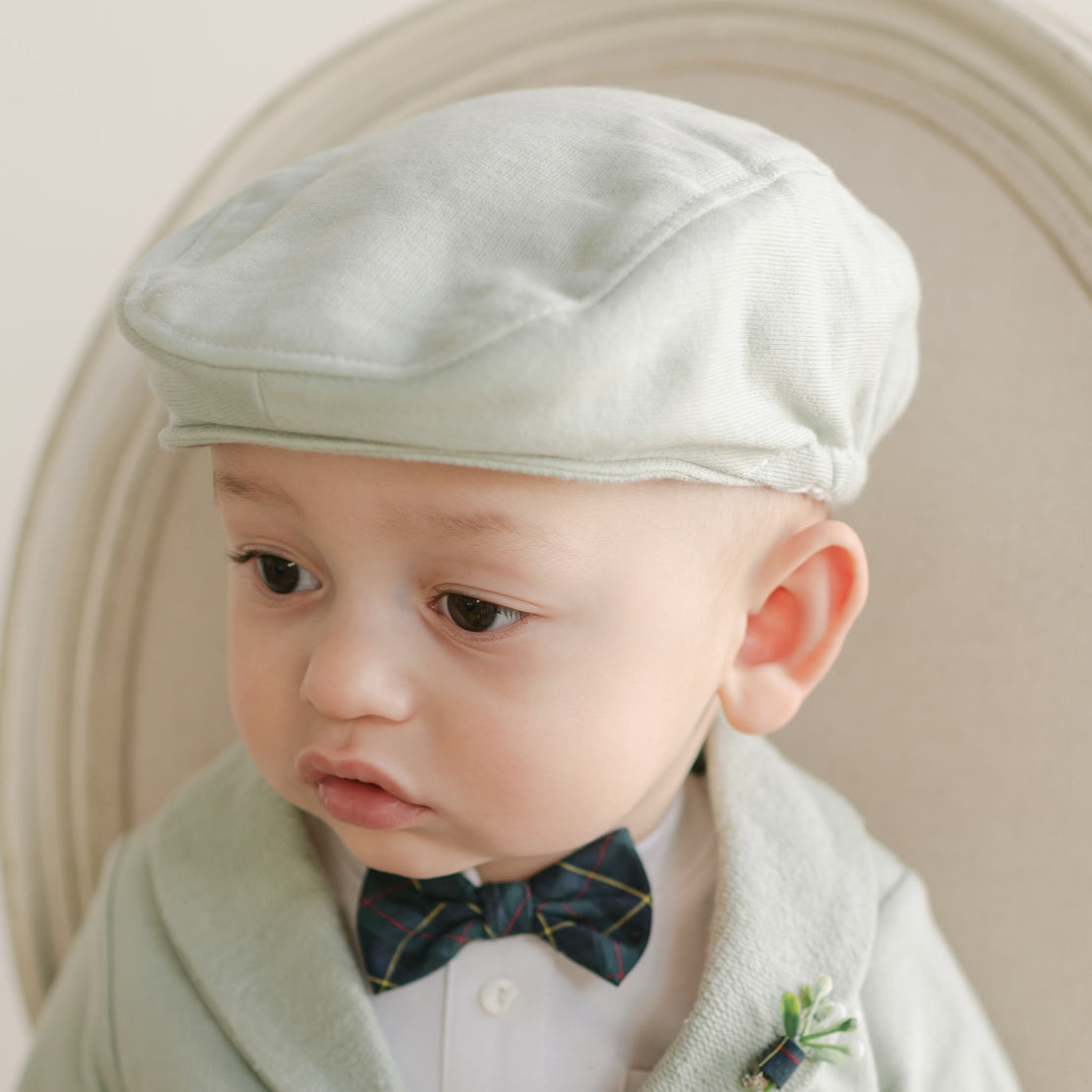 A baby, sporting the Cole Newsboy Cap in light green, along with a matching shawl made from soft French terry cotton, sits in a light-colored chair. Wearing a white shirt and a dark plaid bow tie, the little one gazes to the side with a curious expression.