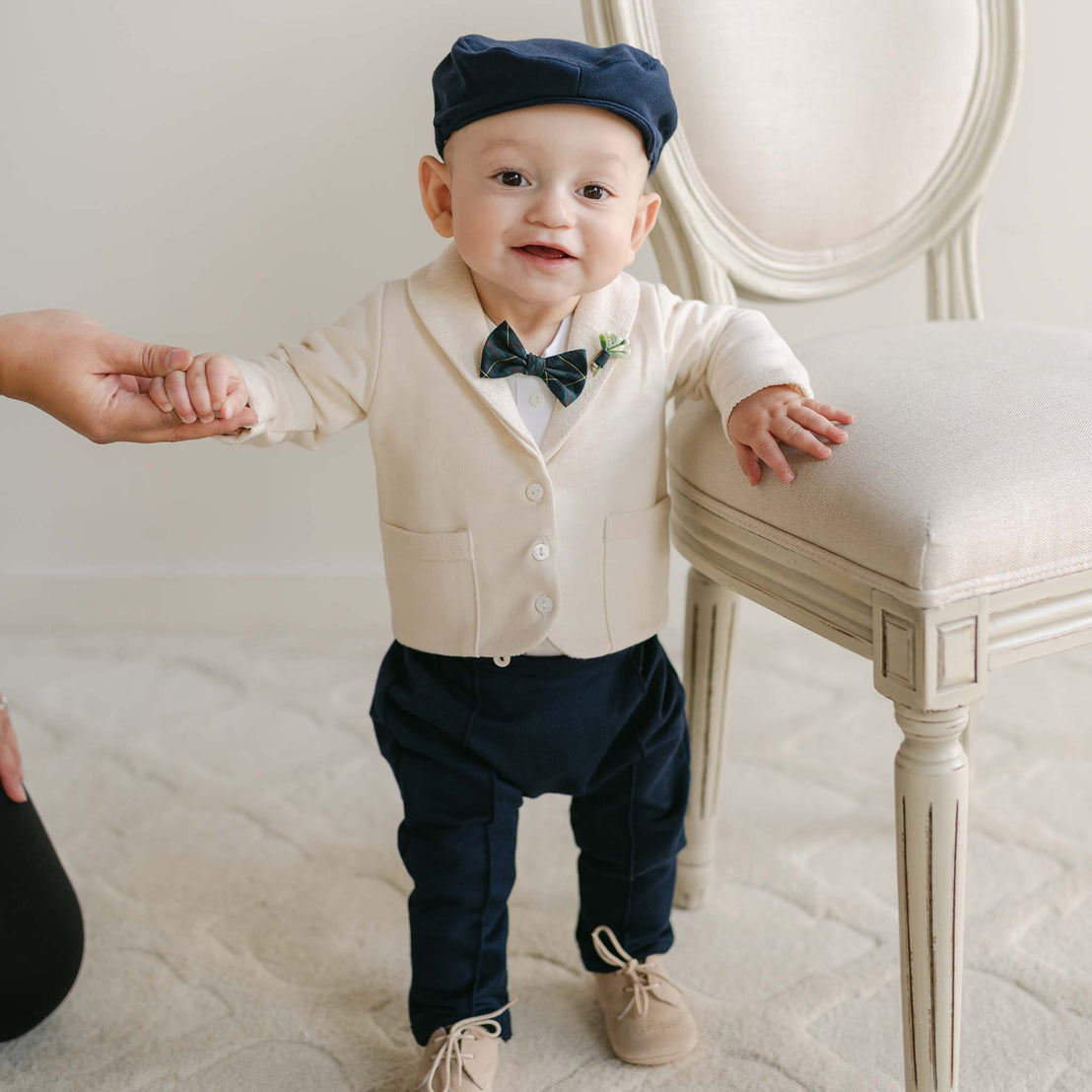 A baby in a Cole 3-Piece Suit, featuring a cream-colored French Terry cotton jacket with a bow tie and blue pants, stands with support from an adult's hand. The baby is smiling, ready for the upcoming holiday party, and there’s a chair nearby.