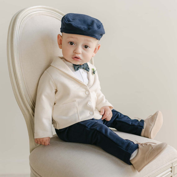 A baby dressed in the Cole Suit & Bow Tie Set, featuring a cream cardigan, dark pants, and a striking blue plaid bow tie, sits on an upholstered chair in a curious pose. The matching cap perfectly completes the outfit, all handmade in the USA.