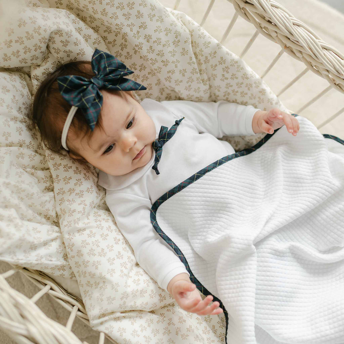 A baby adorned with a Colette Bow Headband on a hand-stretched nylon band is lying in a wicker bassinet. The infant is dressed in a white outfit with a matching bow and covered with a white blanket trimmed in dark plaid. The bassinet is complete with a patterned cushion, enhancing its charm.