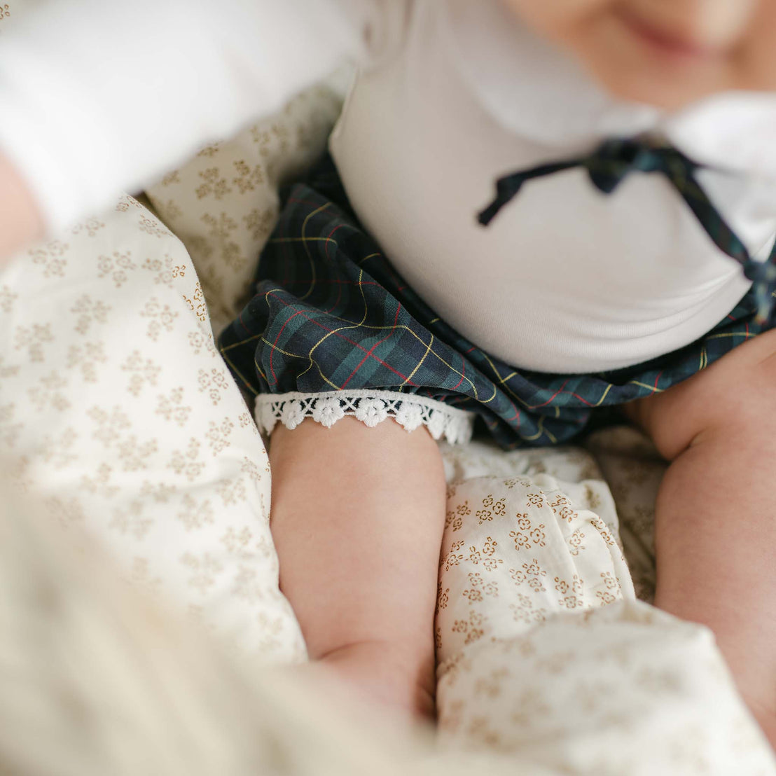 A close-up of a baby sitting on a patterned blanket, wearing the adorable Colette Bloomer Set, which features a charming blue plaid bloomer and onesie with lace trim. The white top beautifully highlights the baby's chubby legs, making it ideal for capturing precious memories at holiday events.