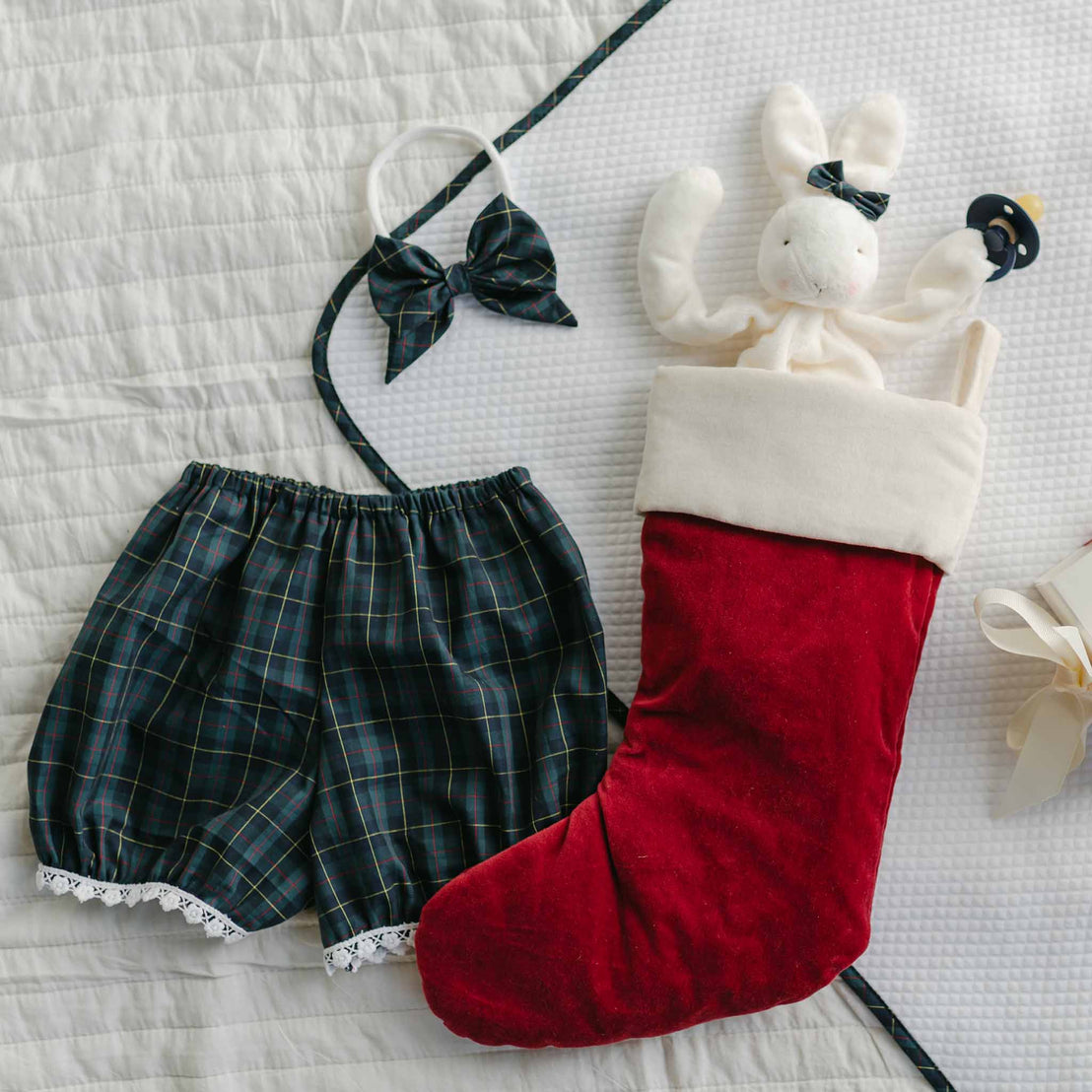 A plaid pair of shorts with lace trim and a matching headband featuring a bow are spread out alongside a red Christmas stocking containing a stuffed bunny toy on a white quilted background. Adding to the charming scene, the Cole Silly Bunny Buddy Pacifier Holder dangles playfully nearby.