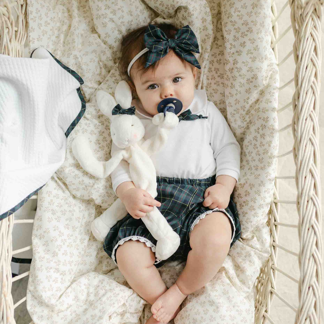 A baby dressed in a Colette Bloomer Set, complete with a plaid outfit and headband, lies in a wicker bassinet, clutching a stuffed rabbit. The baby has a pacifier in their mouth, while a white blanket nearby enhances the serene charm of holiday events amidst cozy surroundings.