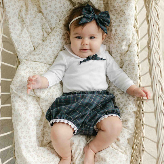 A baby is lying in a woven basket, wearing the Colette Bloomer Set that features a charming white long-sleeve shirt and blue plaid shorts. The little one also sports a matching plaid bow, creating an adorable look perfect for any holiday events. The basket is lined with a patterned cushion, completing this delightful scene.