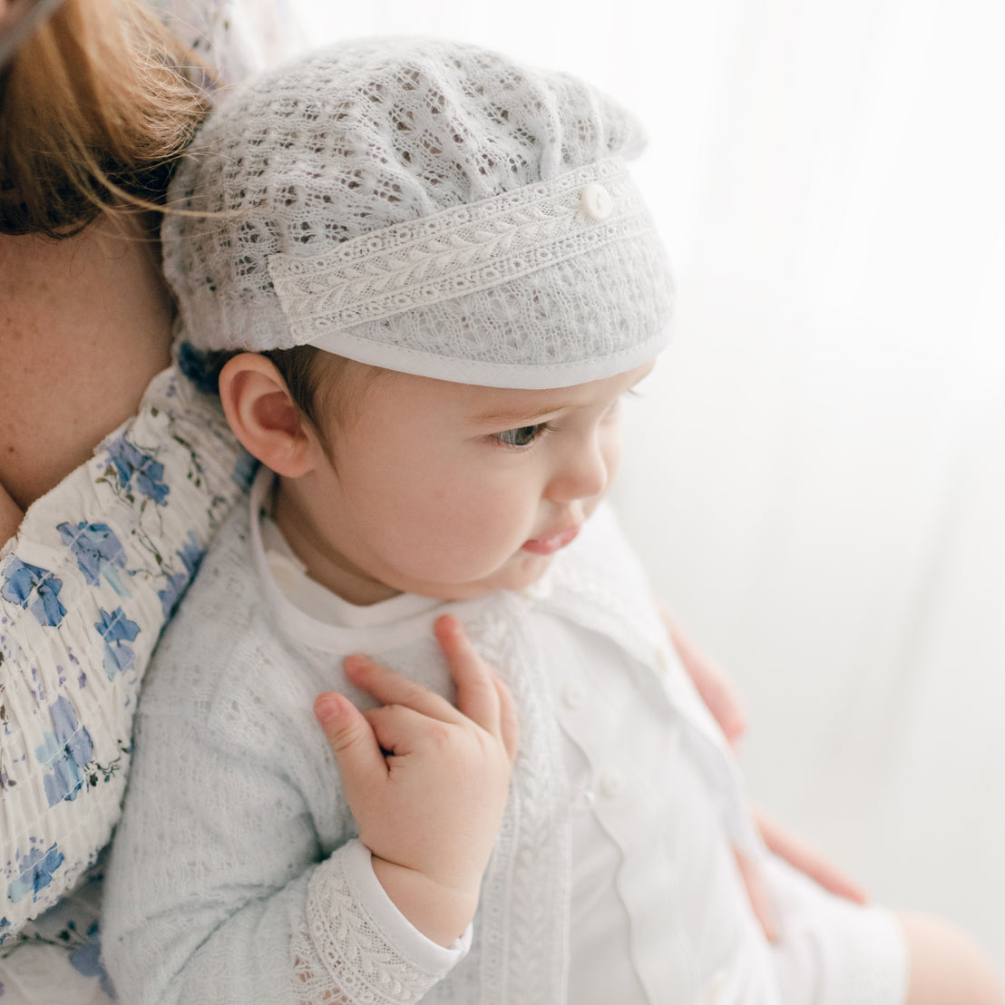 A baby dressed in the Oliver Romper and Knit Sweater and Cap ensemble in white featuring intricate embroidery. He is cradled by a partially visible adult, while he gazes to the side as light streams through a window in the background.