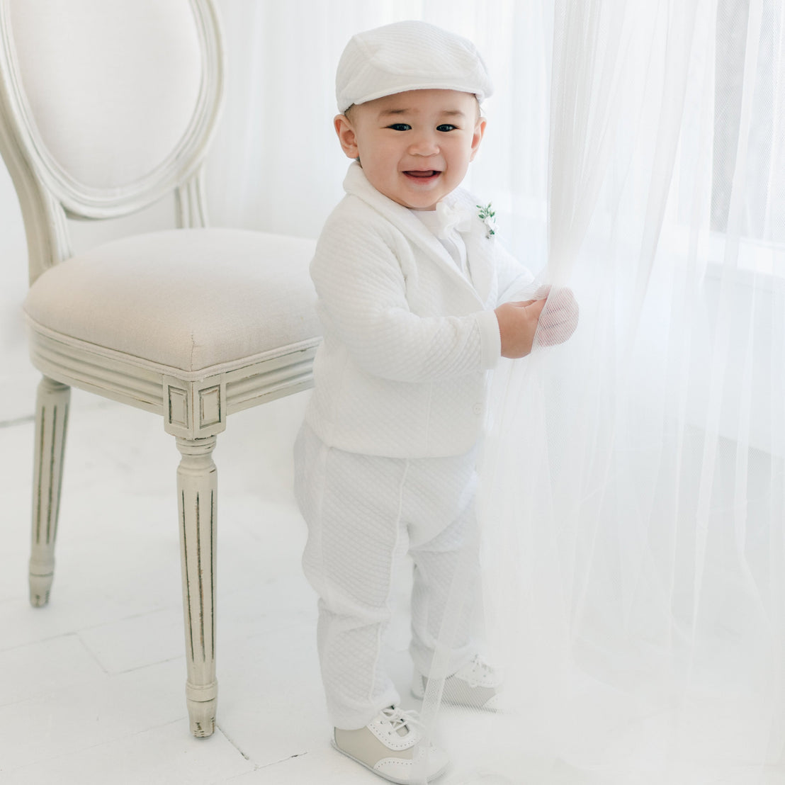 A smiling toddler dressed in an Elijah 3-Piece Suit, including pants, a jacket, and ones is standing near a window. The baby is also wearing a White Velvet Bow Tie & Boutonniere and a matching Elijah Newsboy Cap. is holding a sheer white curtain with one hand and is next to a classic beige upholstered chair. The room has a bright and airy feel.