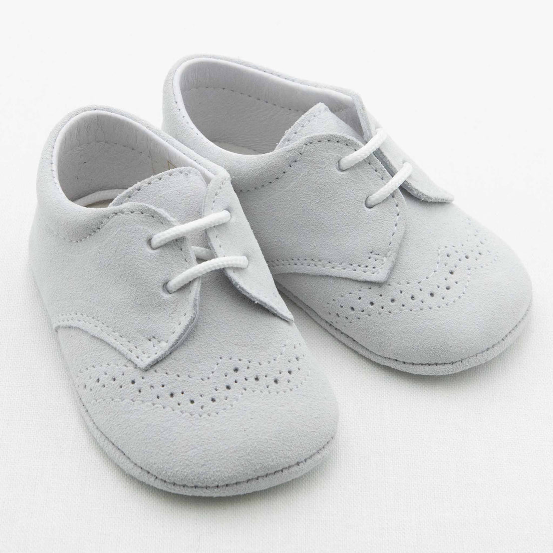 A pair of light gray Ezra Suede Shoes by Baby Beau & Belle is displayed, featuring white laces, a soft suede-like material, decorative perforations on the toe area, and stitching details. These handmade shoes offer a classic and elegant look and are placed on a plain white background.