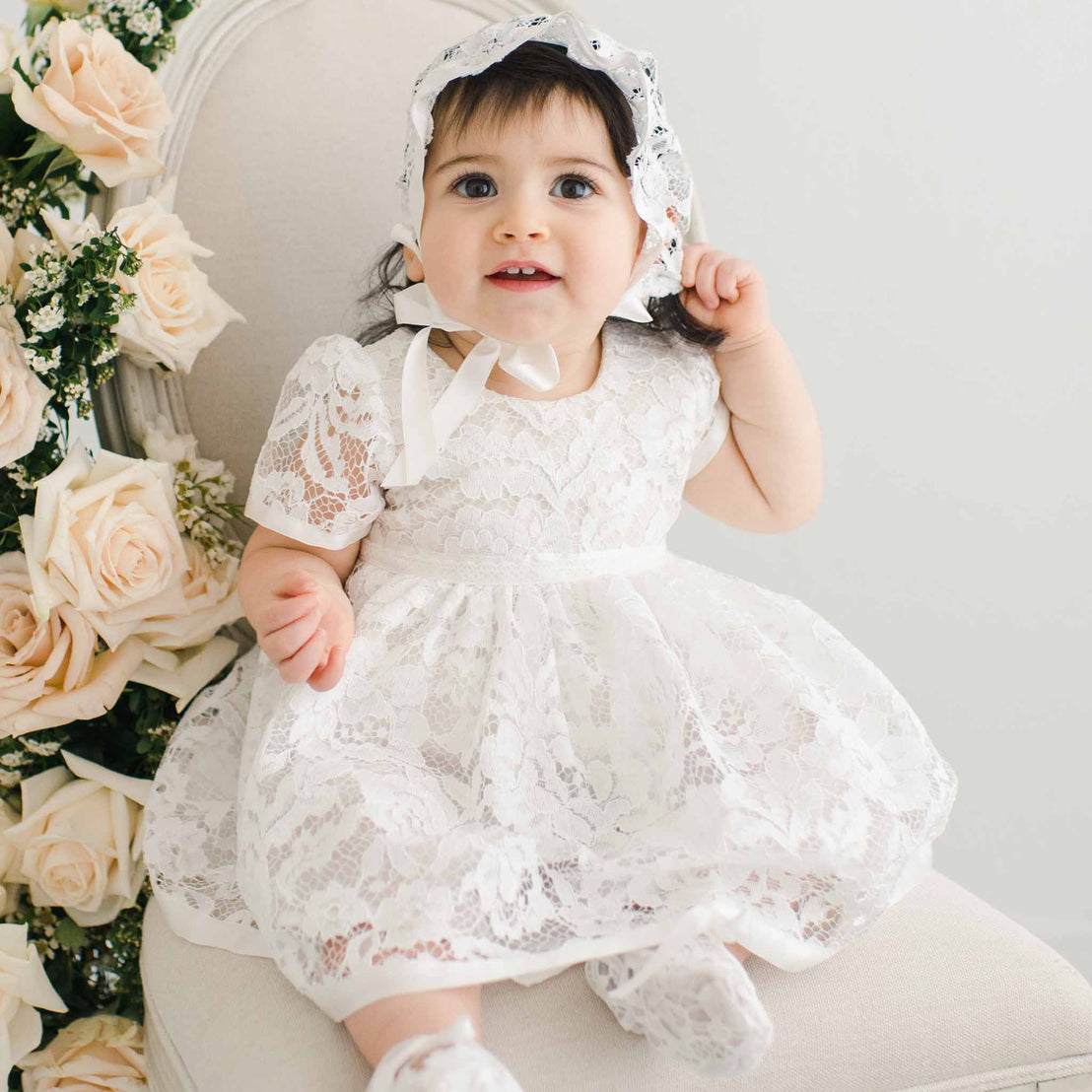 A young child wearing a white lace Rose Romper Dress and bonnet sits on a cream chair, surrounded by pink roses, looking upward with a joyful expression.