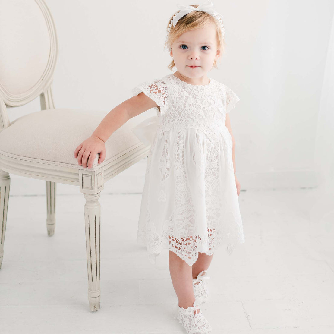 A young girl in a white lace Grace Christening dress and headband stands next to a chair in a softly lit room, looking at the camera with a gentle expression.