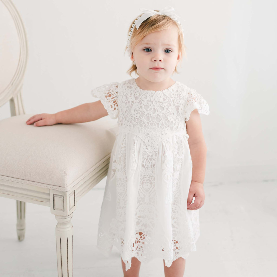 A young child in a Grace Dress standing next to a light-colored chair in a bright room, looking directly at the camera with a subtle expression.