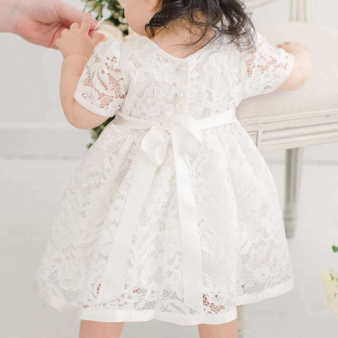 A toddler wearing a Rose Romper Dress, reaching up towards an adult in an upscale, bright, elegant room. Only the child's back is visible.