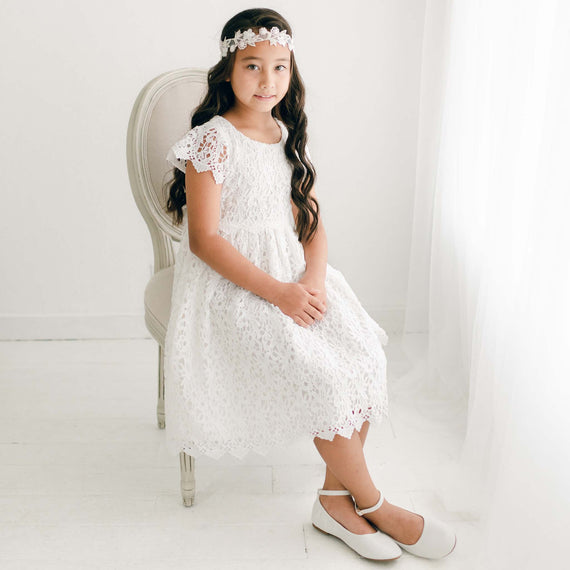 A young girl wearing the Lola Girls Lace Dress in light ivory, featuring intricate all-over embroidered lace, lace cap sleeves, a lace bodice, and button-back closure.