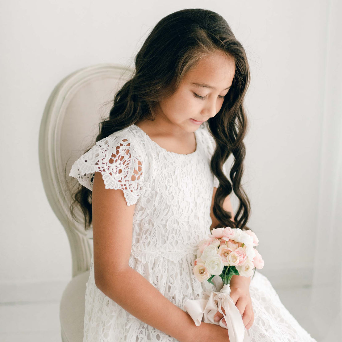 Young girl wearing the Lola Girls Lace Dress, featuring elegant cap sleeves and an all-over lace design. She is seated with a bouquet of pastel flowers, highlighting the dress's intricate lace pattern and soft ivory color.