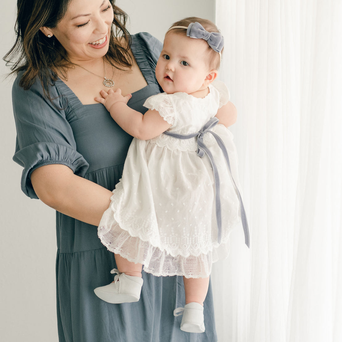 Baby girl wearing the heather Emily Dress, Bloomers, Sash Tie and Emily Velvet Bow Headband. She is being held by her mother and photo shows details of the back of the dress.