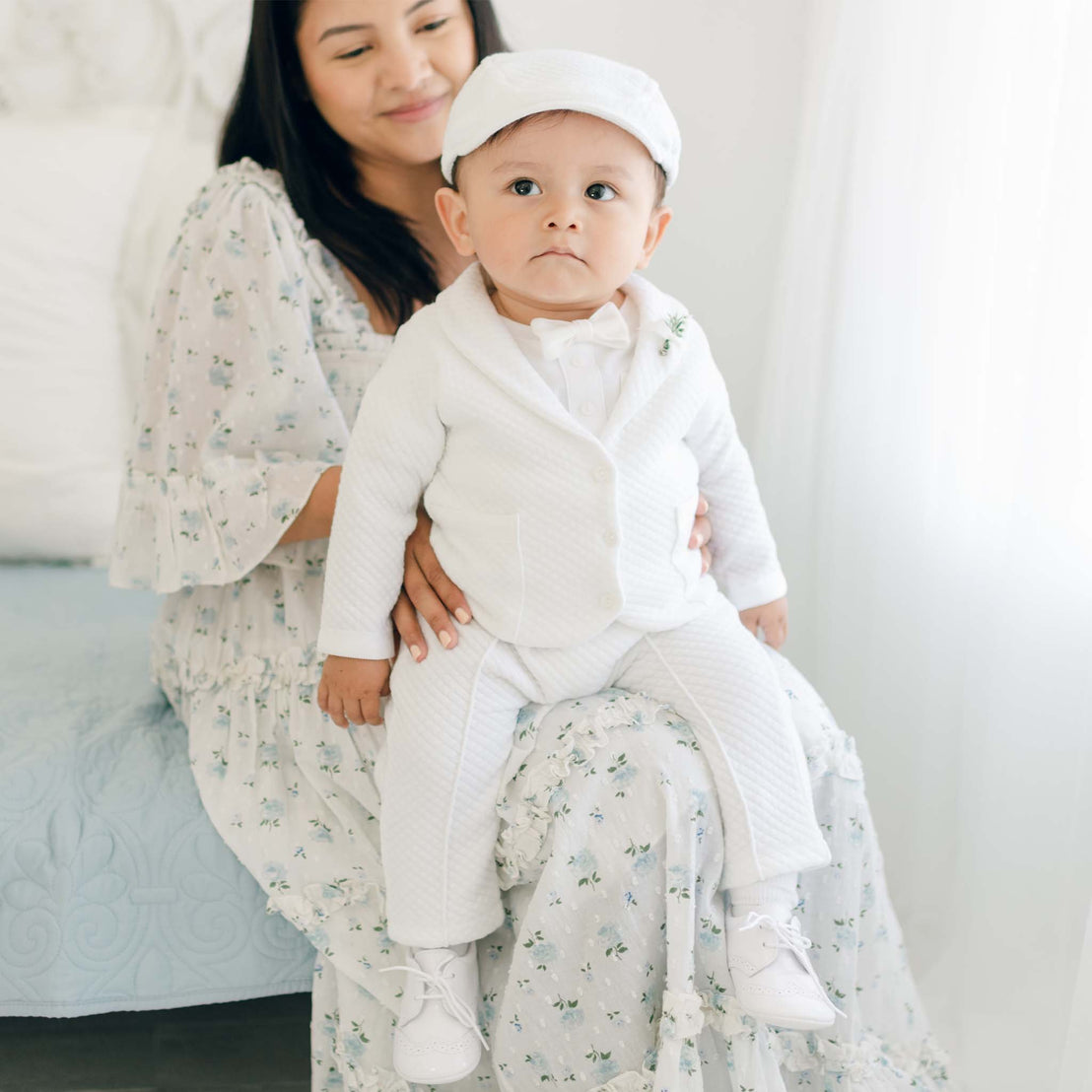 A toddler dressed in the Elijah 3-Piece Suit, complete with a matching cap, sits on a woman's lap. The woman is wearing a floral dress and smiles softly while looking at the child. They are indoors with a light-filled background, capturing the essence of this special occasion.