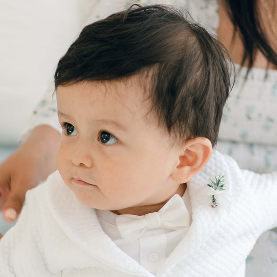 A young child with dark hair, dressed in a white outfit featuring a White Velvet Bow Tie & Boutonniere, looks to the side. The ensemble includes a white shirt, jacket adorned with the small decorative pin on the lapel, and the elegant bow tie. An adult is partially visible in the background, wearing a floral patterned top.