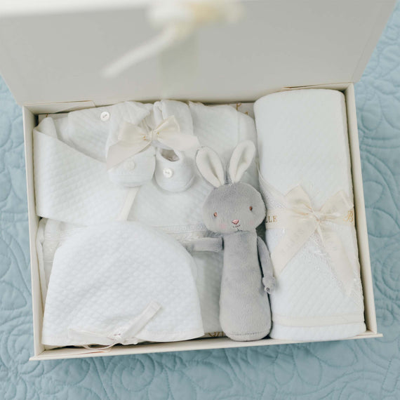 A neatly arranged baby gift box contains a soft gray bunny toy, a white quilted baby blanket, a matching hat, and an adorable baby gown tied with cream-colored ribbons – the perfect Elijah Newborn Gift Set - Save 10% for a delightful baby shower gift. All items rest on a pale blue quilted surface.