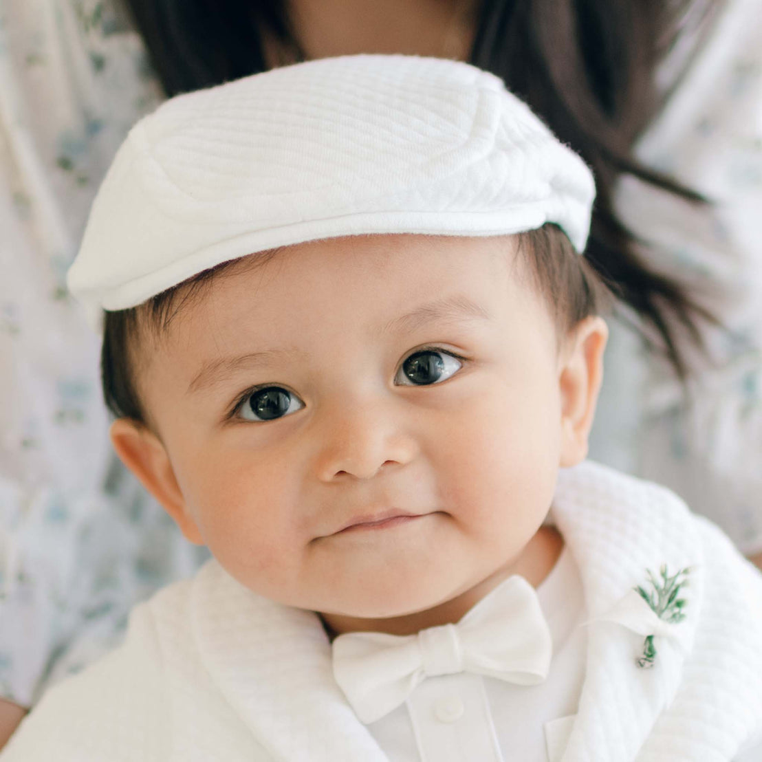 A baby dressed in a handmade, textured cotton outfit with the Elijah Newsboy Cap and a matching bow tie looks at the camera with a slight smile. The background is softly blurred, and an adult appears to be holding the baby.