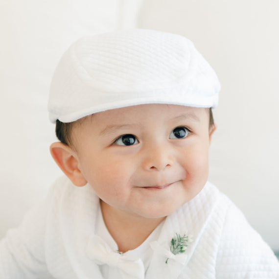 A baby wearing the Elijah Newsboy Cap and a textured cotton outfit looks slightly to the side with a smile. The baby also has a small sprig of greenery pinned to their outfit near the bow tie. The background is a soft white, highlighting the handmade details of their charming, newsboy style ensemble.