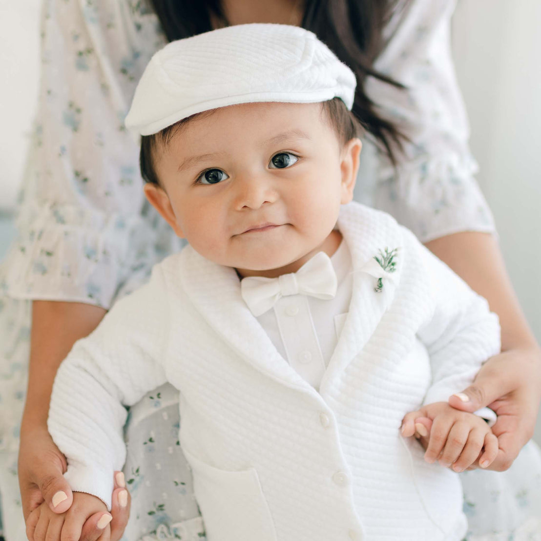 A baby dressed in the Elijah 3-Piece Suit, complete with a bow tie and hat, is being gently held and supported by an adult partially visible in the background. The baby smiles at the camera with large, bright eyes, exuding a sense of curiosity and sweetness perfect for any special occasion.