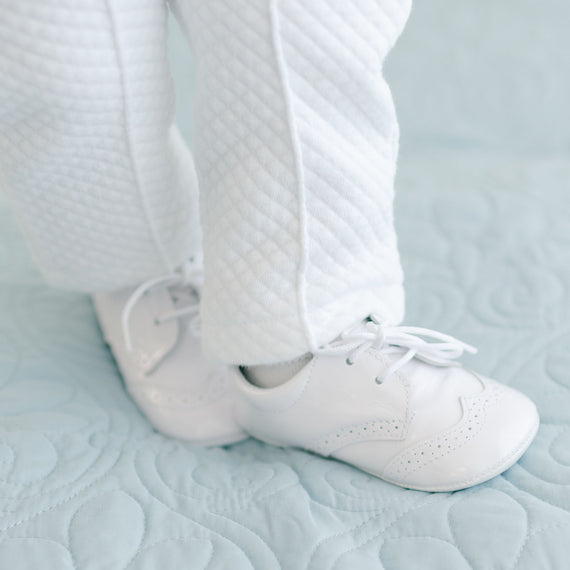 A close-up of a baby's feet wearing the White Patent Leather Shoes and Elijah White Quilt Pants, standing on a light blue quilted blanket.