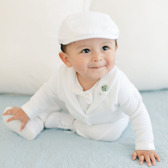 A baby boy in the Elijah 3-Piece Suit and cap is sitting on a light blue blanket. The outfit, perfect for a special occasion, features textured cotton. He has a slight smile and is looking slightly upward, with white pillows in the background.