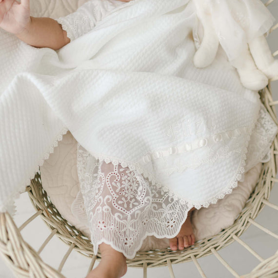 A baby, partially covered by an Eliza Personalized Blanket, rests in a wicker basket. The baby is wearing a delicate white lace outfit, with small feet peeking out. A plush toy can be seen near the baby's head.