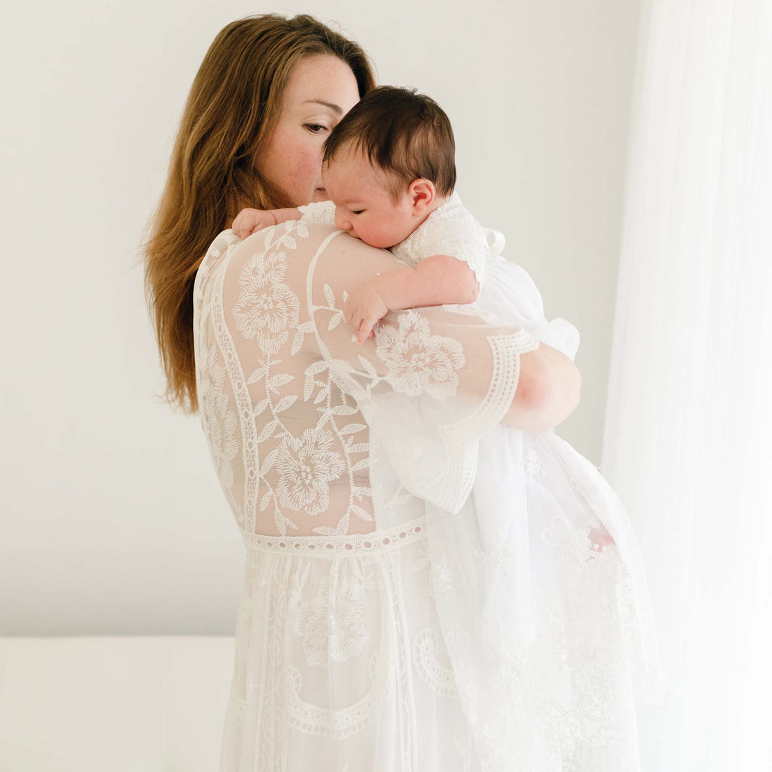 A mother gently holds and kisses her sleeping newborn baby, both dressed in the Eliza Blessing Gown & Bonnet for a christening, against a soft white background.